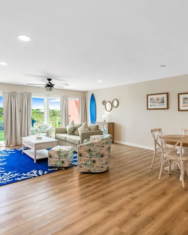 Open-plan living and dining area with beige walls, wood flooring, coastal decor, and glass doors opening to green landscape views