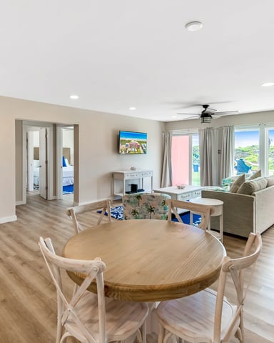Open-plan living and dining area with round wood table, beige sofa, large windows overlooking hillside landscape, and recessed lighting.