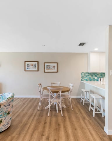 Open-plan living room with dining area, blue surfboard, coastal-themed furniture, and kitchenette with turquoise tile backsplash