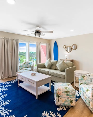 Bright living room with beige sofa, blue coral-patterned rug, coastal décor, and glass doors opening to hillside views