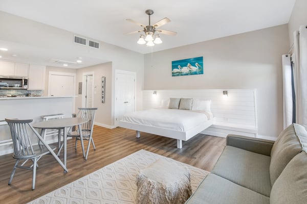 Open studio apartment with white bed, dining table, kitchen, and beige sofa on wood flooring