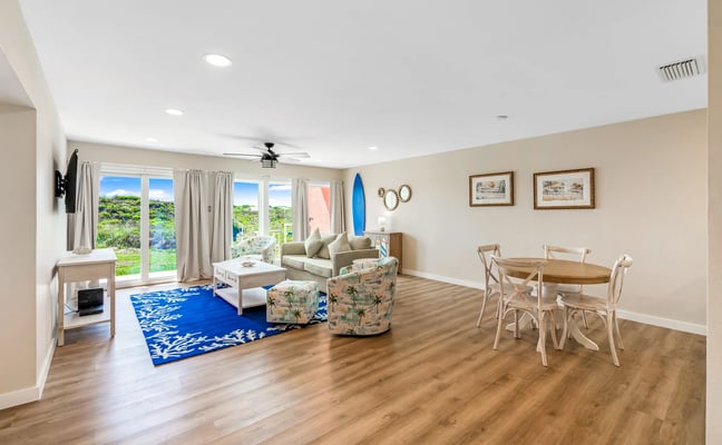 Open-plan living and dining room with beige walls, wood flooring, glass doors opening to green landscape, and coastal-themed decor including surfboard and blue