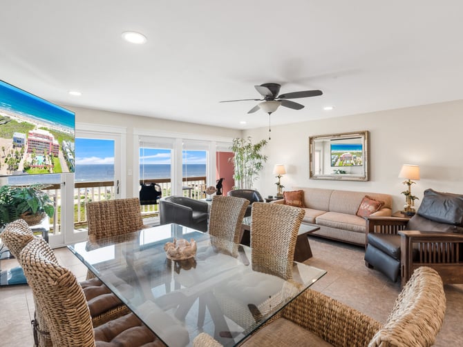 Open-plan living room with ocean view, wicker dining chairs, glass table, beige sofa, and coastal decor