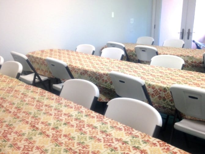 Long tables covered with patterned fabric and white chairs arranged for a meeting or event in a bright room with glass doors.