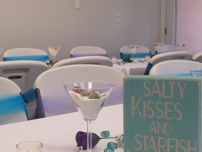 Banquet table with white linens, cocktail glass, candle, turquoise decorative sign reading "Salty Kisses and Starfish Wishes", and white chairs with blue