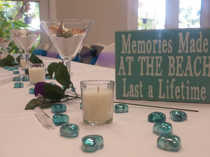 Decorated dining table with martini glasses, candles, purple flowers, turquoise stones, and green sign reading "Memories Made at the Beach Last a Lifetime"